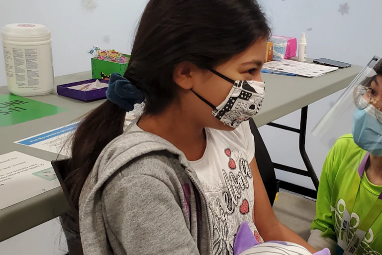 A girl wearing a mask with her hair in a ponytail about to get her COVID-19 vaccination.