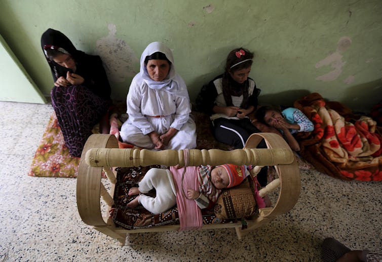 Three women sitting on a carpet with a child on a cot in front of them.