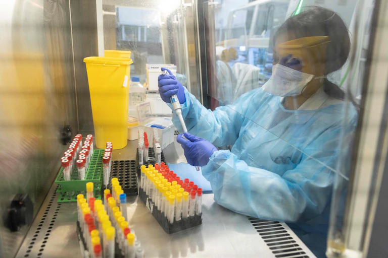 masked researcher in PPE works with test tubes under a protective hood