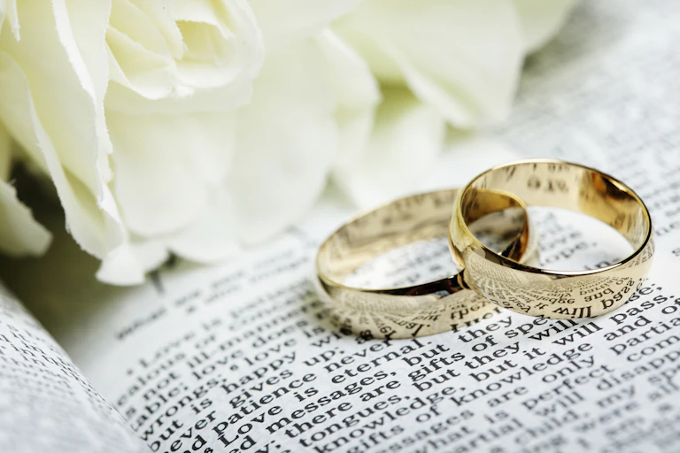 Two wedding rings sit on an open Bible, next to white flowers.