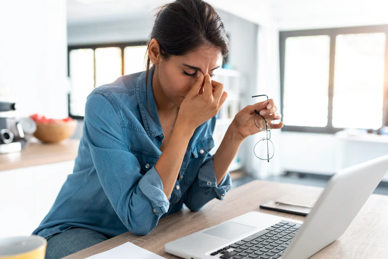 A woman seated in front of a laptop computer with her eyes closed, her right hand pinching the bridge of her nose, her left hand holding a pair of eye glasses