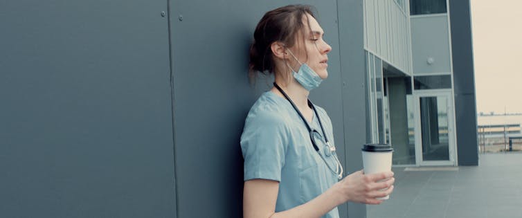 Exhausted health worker leaning on hospital wall holding cup of coffee