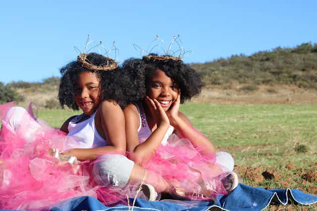 Twin girls in pink skirts and crowns sit, smiling.