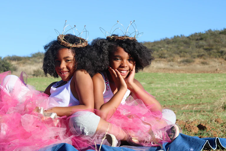 Twin girls in pink skirts and crowns sit, smiling.