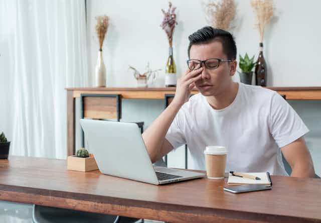 An Asian man sits behind his laptop with his hand over his face looking exhausted