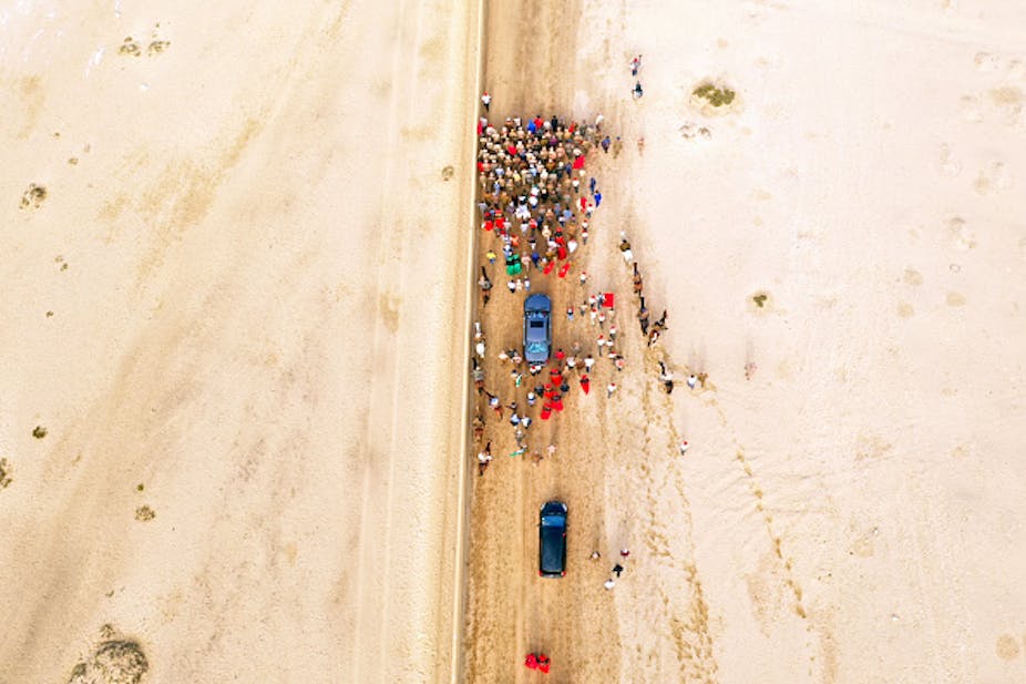 From above, a small crowd of brightly dressed people walking down a sand road in a desert, two cars trailing them.