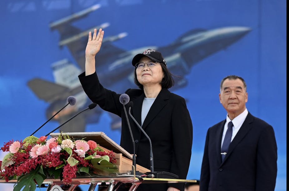 La présidente taïwanaise salue à un pupitre fleuri. Derrière elle, une image d'avion de chasse.