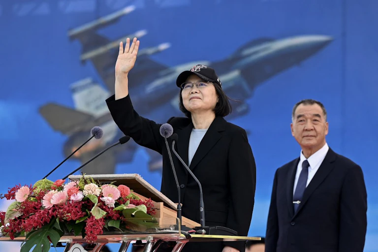 La présidente taïwanaise salue à un pupitre fleuri. Derrière elle, une image d'avion de chasse.