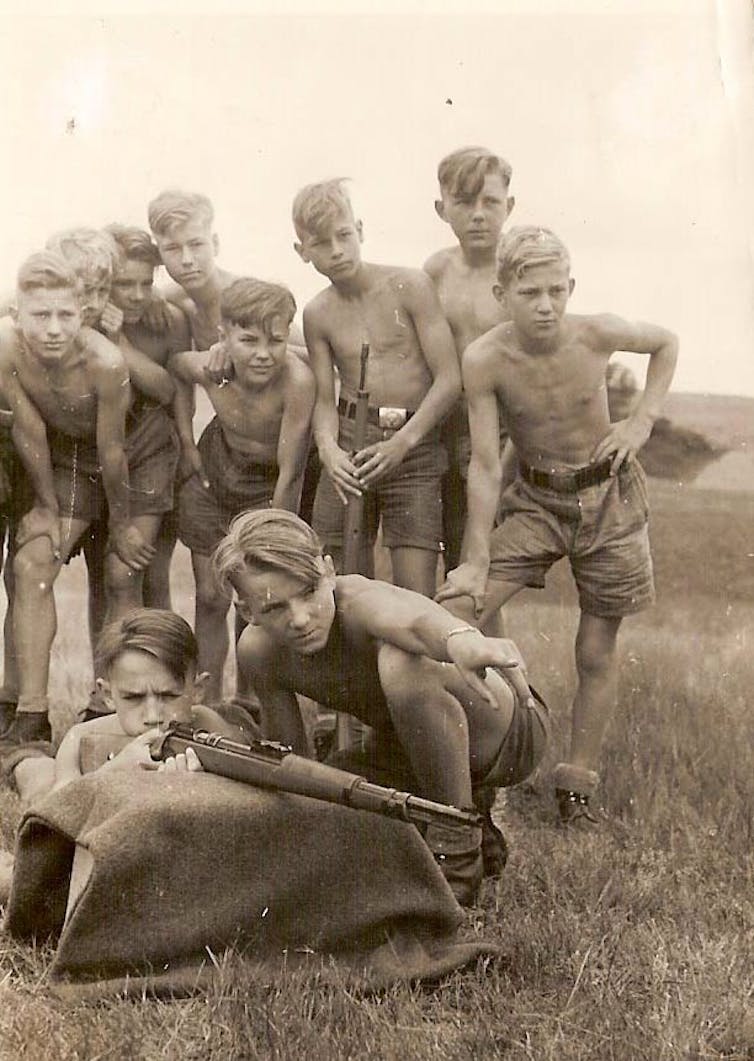 Almost a dozen teen boys, shirtless, gathered around a fellow student who is aiming a rifle at something.