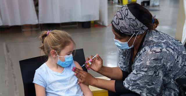 A girl with a ponytail, blue mask and blue t-shirt looks at her arm as a health-care worker vaccinates her