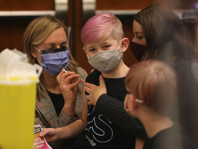 A boy with pink hair and wearing a mask looks away from a health-care worker as she vaccinates him.