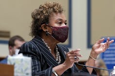 A woman wearing a mask and in a suit and pearls talks into a microphone.