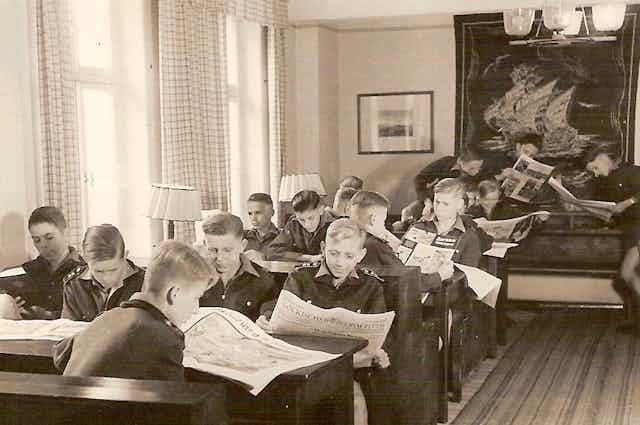 Teen boys in a reading room, sitting at tables reading newspapers. 