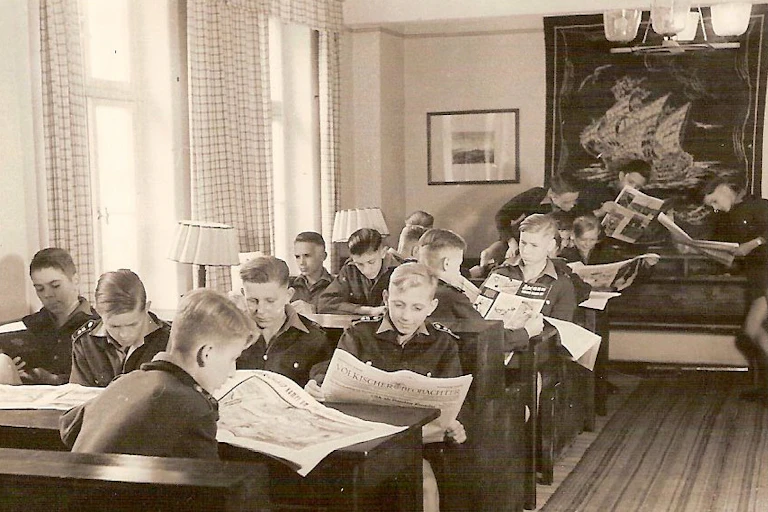 Teen boys in a reading room, sitting at tables reading newspapers.