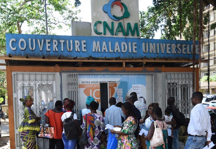 File d'attente devant un petit bâtiment sur lequel il est écrit Couverture Maladie Universelle.