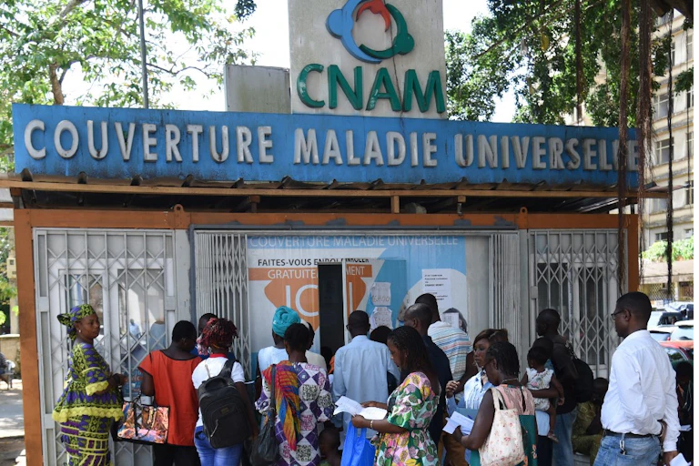 File d'attente devant un petit bâtiment sur lequel il est écrit "Couverture Maladie Universelle".