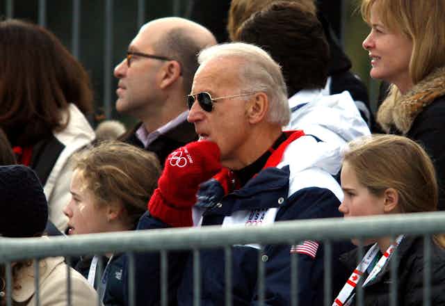 Joe Biden sits wearing a team USA jacket, olympic mits and aviator sunglasses