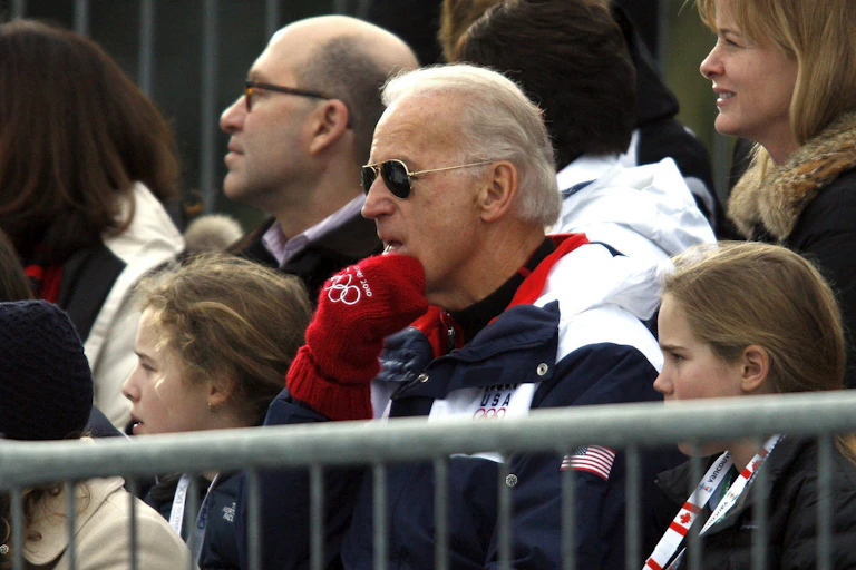 Joe Biden sits wearing a team USA jacket, olympic mits and aviator sunglasses