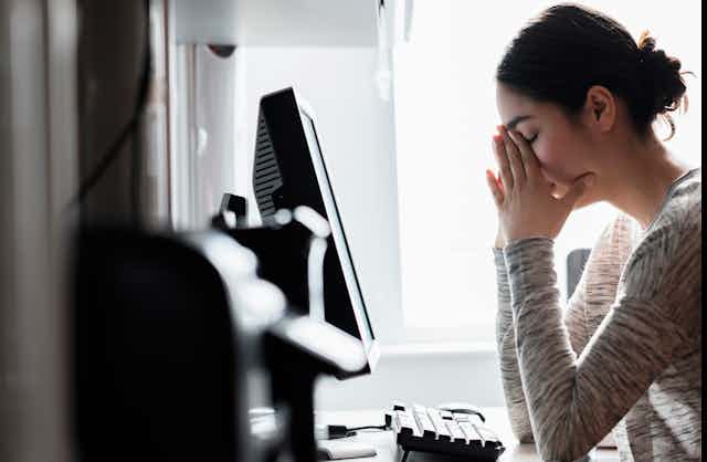 An anxiety-filled young woman, sitting in front of her computer screen, covers her face with her hands.