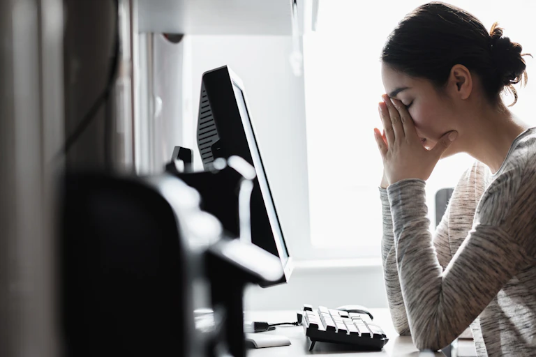 An anxiety-filled young woman, sitting in front of her computer screen, covers her face with her hands.