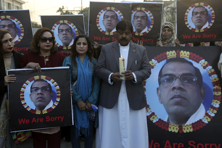 A man dressed in a jacket and white long shirt holding a candle, while two women standing next to home hold the photograph of a man, who was lynched in Pakistan.