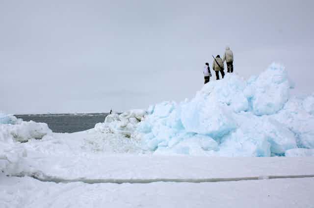 Three people stand on a pile of ice and snow at the edge of the ocean.