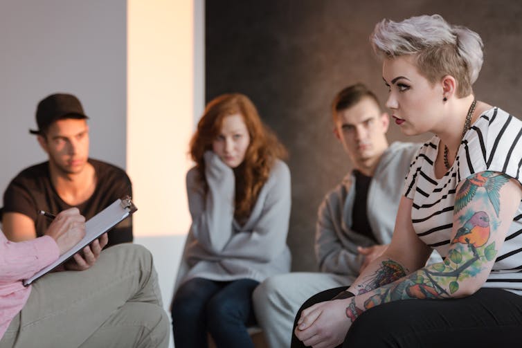 A tattooed teenage girl speaks to a therapist during a group psychotherapy meeting. Three other young people, sitting in the background, are listening.