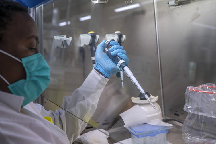 A woman in a lab coat, face mask and gloves using a pipette in a lab