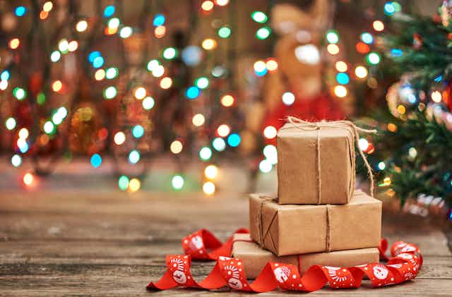 A stack of three gifts wrapped in brown paper, with a tree and twinkling lights in the background