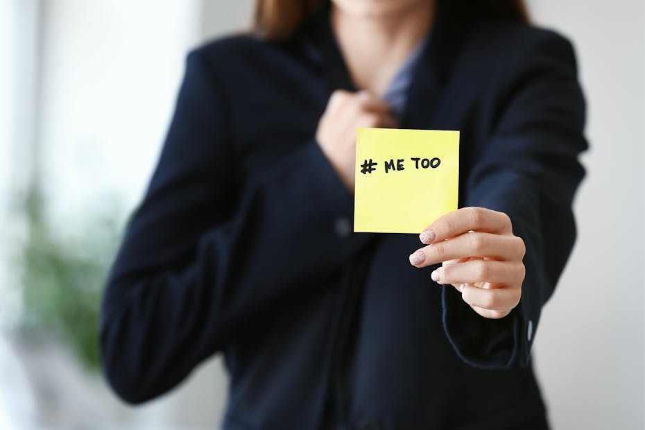A woman hold up a post-it that says 'me too.'