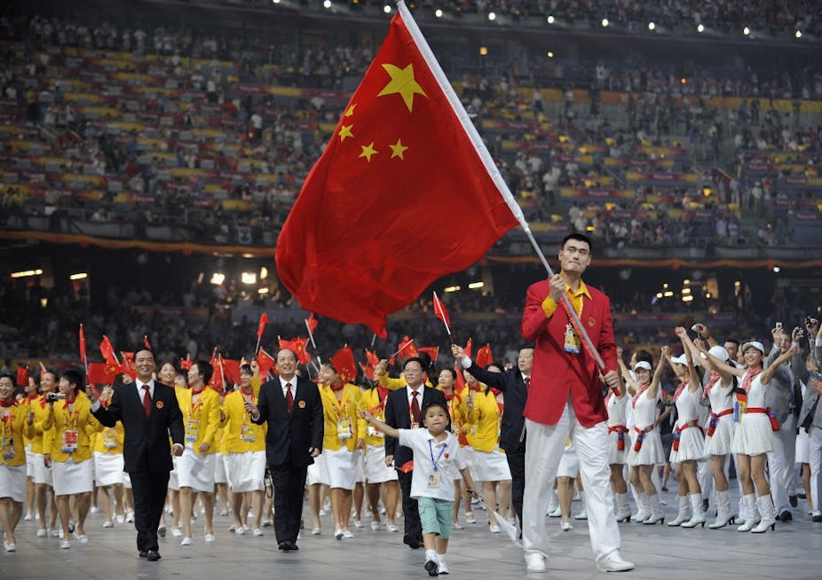 Un homme porte le drapeau chinois dans un stade, suivi par une foule de personnes agitant des drapeaux chinois miniatures.