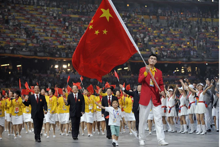 Un homme porte le drapeau chinois dans un stade, suivi par une foule de personnes agitant des drapeaux chinois miniatures.