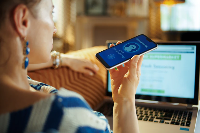 A woman holds a phone up near a computer screen.