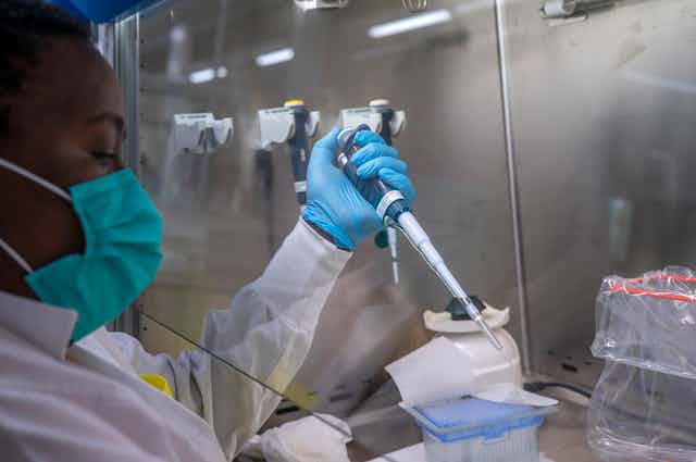 A person wearing a facemask, lab coat and rubber gloves holds a long syringe-like device 