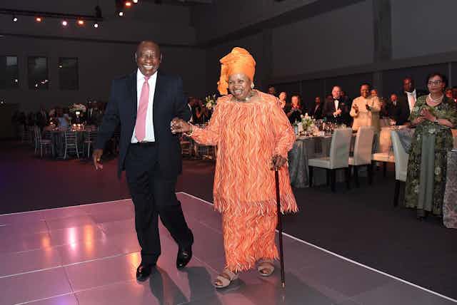 A woman, dressed in bright orange traditional attire and carrying a walking stick, laughs joyfully as she is accompanied by a man in a suit onto a stage. All around people stand and applaud.