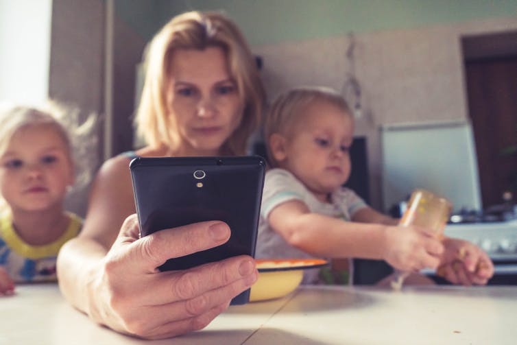 Mother organises things on her phone, while looking after kids at the kitchen table.