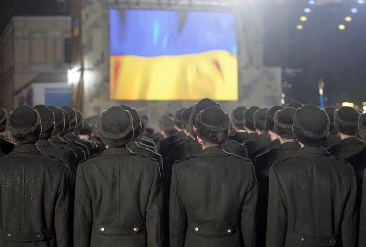 Soldiers stand in line, with their backs to the camera, in front of a Ukrainian flag.