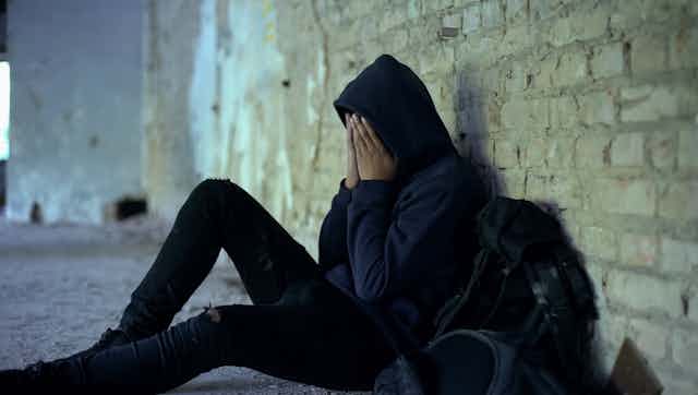 A teenager sits on the ground, leaned up against a brick wall with their hands on their face.