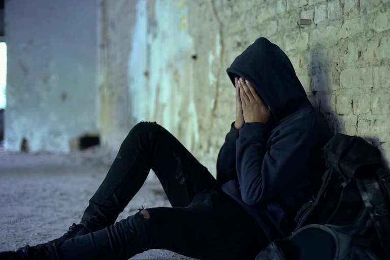 A teenager sits on the ground, leaned up against a brick wall with their hands on their face.
