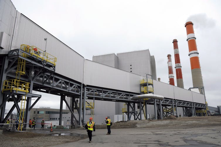 Three orange and white smokestacks in the background rise above a series of industrial buildings and corridors, with a few people in safety vests walking around.