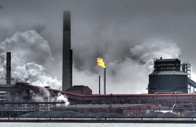 A flare burns off gas from a stack, surrounded by other stacks and plumes of smoke and gases. More than a dozen piles of coal sit in front of the buildings.