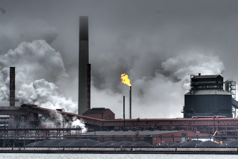 A flare burns off gas from a stack, surrounded by other stacks and plumes of smoke and gases. More than a dozen piles of coal sit in front of the buildings.