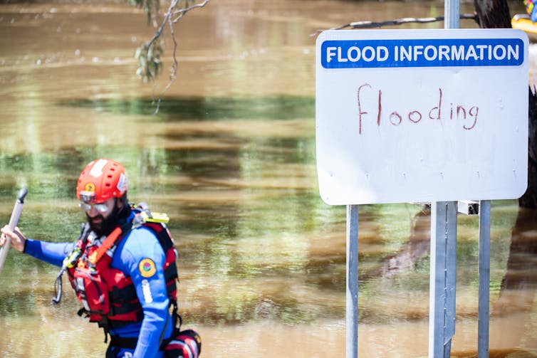man in front of flood waters and flood warning sign