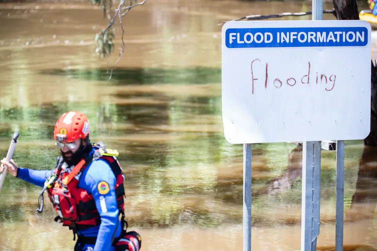 man in front of flood waters and flood warning sign