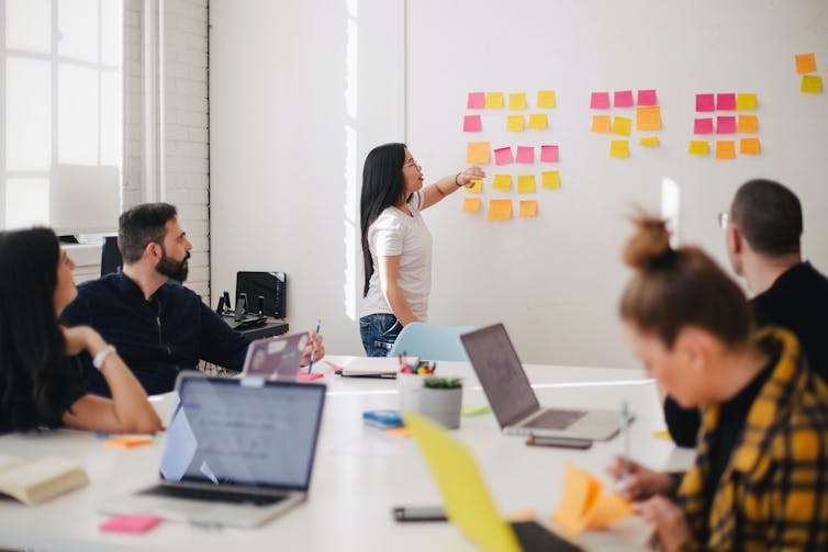 Young people in a boardroom put sticky notes on a white board.