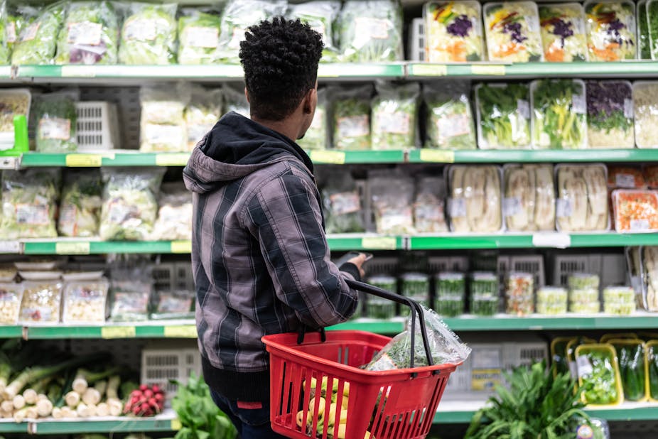 A young man holding a red basket looks at rows of fresh produce displayed on supermarket shelves.