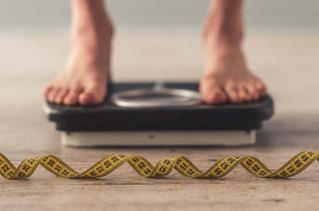 Feet standing on weigh scales in a blurred background, while a tape measure lays on the floor in the foreground in focus