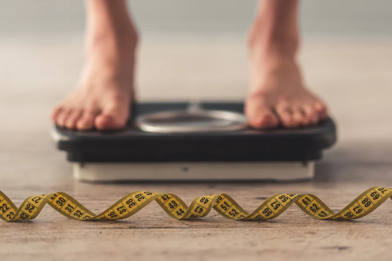 Feet standing on weigh scales in a blurred background, while a tape measure lays on the floor in the foreground in focus