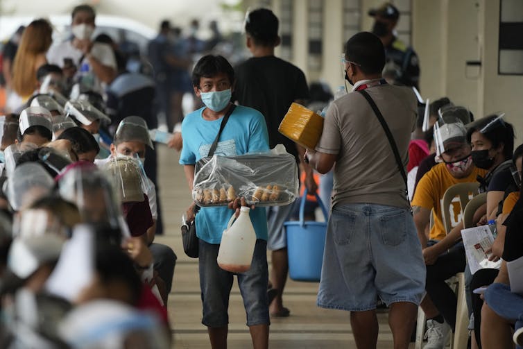 A person walks up and down rows of people selling food as they wait in line for vaccines.