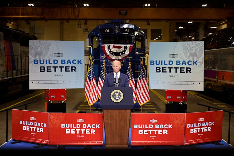 Joe Biden habla en un atril frente a grandes carteles de Building Back Better.  Banderas estadounidenses lo flanquean en el podio.